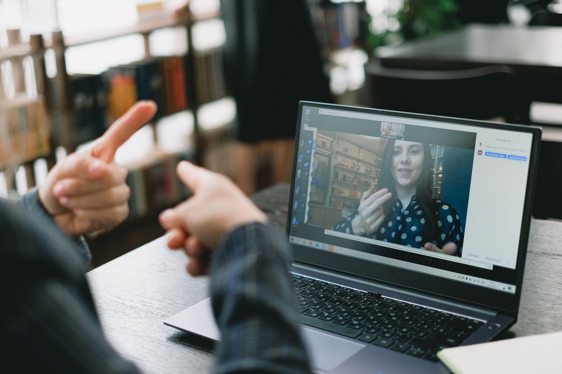 Duas mulheres se comunicam em Libras durante uma videochamada em um notebook, em um ambiente interno iluminado.