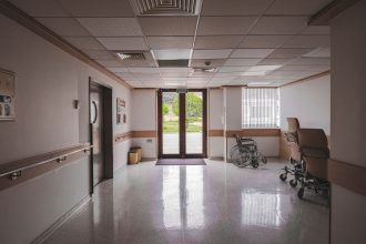 Cadeira de rodas em corredor de hospital, diante de portas de vidro com vista para a área externa