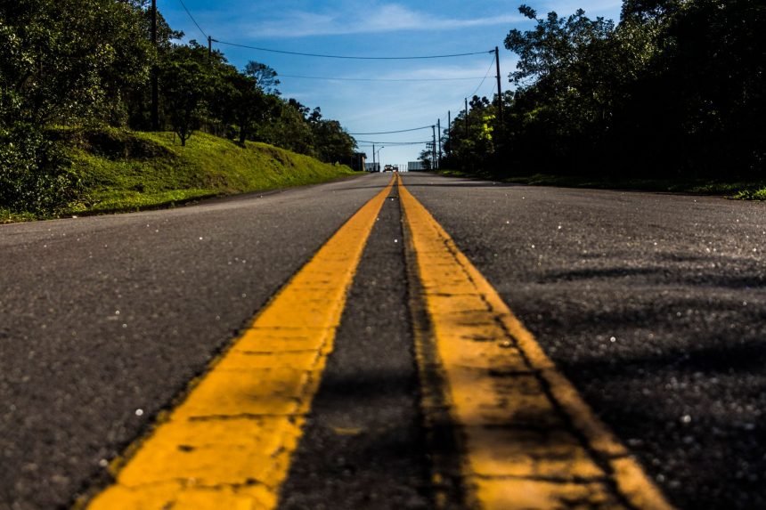 Rodovia pavimentada com faixa amarela, cercada por vegetação sob céu azul