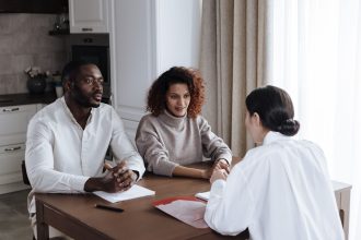 Casal conversa com uma assistente social enquanto analisa documentos em um ambiente residencial.
