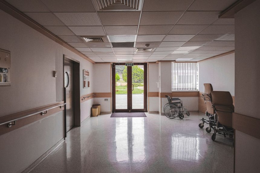 Cadeira de rodas em corredor de hospital, diante de portas de vidro com vista para a área externa