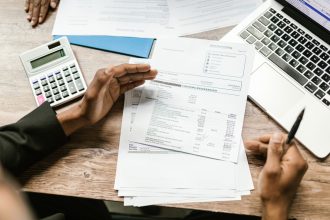 Mãos analisam documentos financeiros ao lado de uma calculadora e um notebook sobre uma mesa.