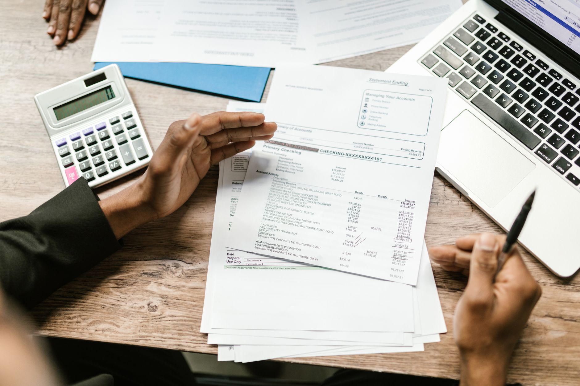 Mãos analisam documentos financeiros ao lado de uma calculadora e um notebook sobre uma mesa.