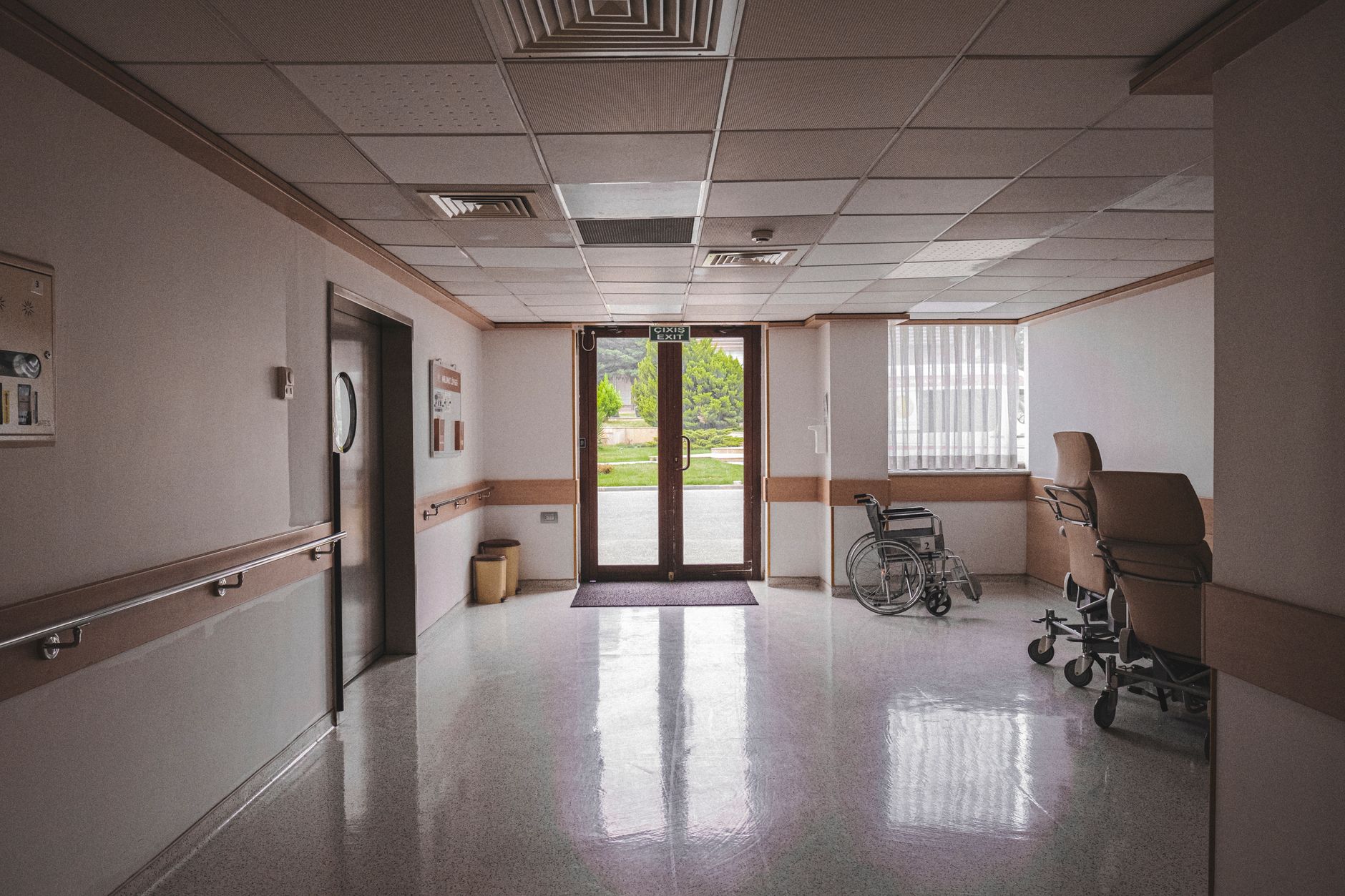 Cadeira de rodas em corredor de hospital, diante de portas de vidro com vista para a área externa