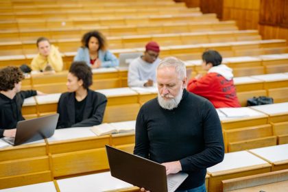 Professor interage com estudantes em uma sala de aula universitária, onde alguns usam laptops.